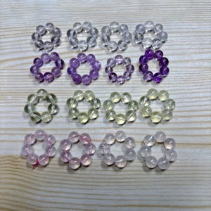 May include: Sixteen circular rings arranged in a four-by-four grid, each constructed from small, smooth, spherical beads. The collection features various translucent colors including clear, purple, light green, and soft pink, all strung together.