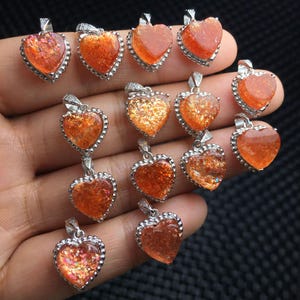 Puede incluir: Una colección de colgantes en forma de corazón con material naranja brillante similar a la piedra solar, engastados en marcos de metal plateado. Cada colgante incluye una pequeña anilla plateada en la parte superior y una superficie texturizada.
