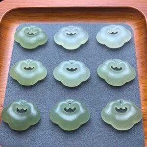 May include: Nine pale green cloud-shaped pendants with a small hole at the top for stringing. The pendants are arranged in a 3x3 grid on a gray surface, set within a wooden tray.