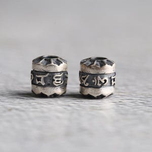 May include: Two cylindrical silver beads featuring a dark oxidized finish and engraved Tibetan script. Each bead has a central band with the text S925 and a decorative sunburst pattern on the top and bottom edges, showing a metallic texture.