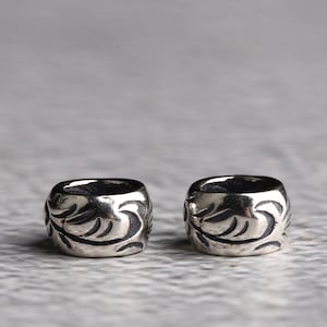 Puede incluir: Dos cuentas cilíndricas plateadas con un diseño floral. Las cuentas tienen una forma cuadrada ligeramente redondeada y un interior oscuro. El patrón floral está grabado en la superficie, creando un contraste.