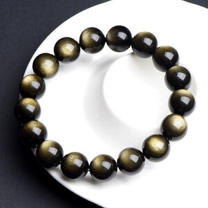 K&ouml;nnte beinhalten: Ein schwarzes und goldenes Perlenarmband mit gl&auml;nzender, irisierender Oberfl&auml;che. Die Perlen sind rund und glatt, und das Armband ist aus Naturstein.