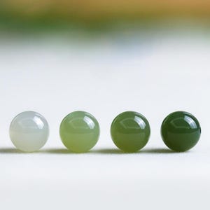 Könnte beinhalten: Vier polierte, runde Edelsteine in verschiedenen Grüntönen. Die Steine gehen von einem blassen, fast weiß-grünen Farbton in ein tiefes, waldgrünes Grün über. Die glatten, reflektierenden Oberflächen deuten auf eine hochwertige Verarbeitung hin.