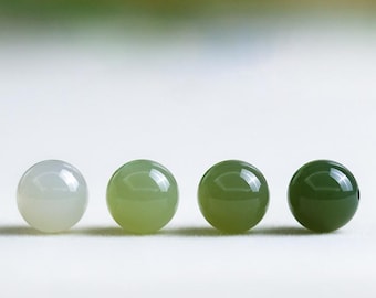 May include: Four polished, round gemstones in varying shades of green. The stones transition from a pale, almost white-green to a deep, forest green. The smooth, reflective surfaces suggest a high-quality finish, ideal for jewelry making.