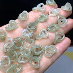May include: A collection of pale green jade heart-shaped charms. The charms are linked together in pairs and scattered across a hand, with a dark background. The translucent jade has a smooth, polished appearance.