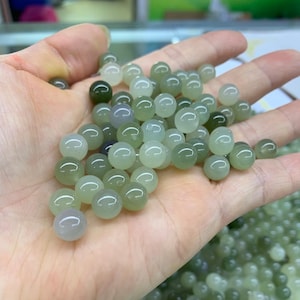 May include: A collection of round, polished beads in shades of green and white. The beads are smooth and appear to be made of stone or glass. The beads are being held in a person's hand. The beads are a light green colour.