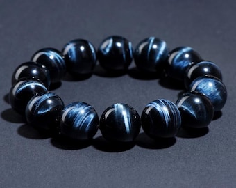 Puede incluir: Un brazalete hecho de cuentas de ojo de tigre azul oscuro. Las cuentas son redondas y tienen un acabado liso y pulido. El brazalete está encordado en un cordón elástico transparente.