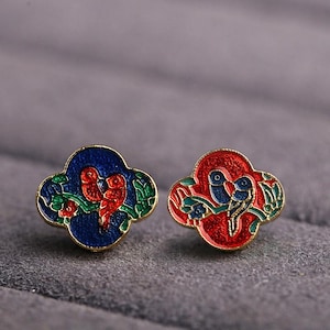 Op de afbeelding: Een paar decoratieve emaille-spelden met een gestileerd bloemmotief met twee vogels. Eén speld heeft rode vogels op een blauwe achtergrond, en de andere heeft blauwe vogels op een rode achtergrond.