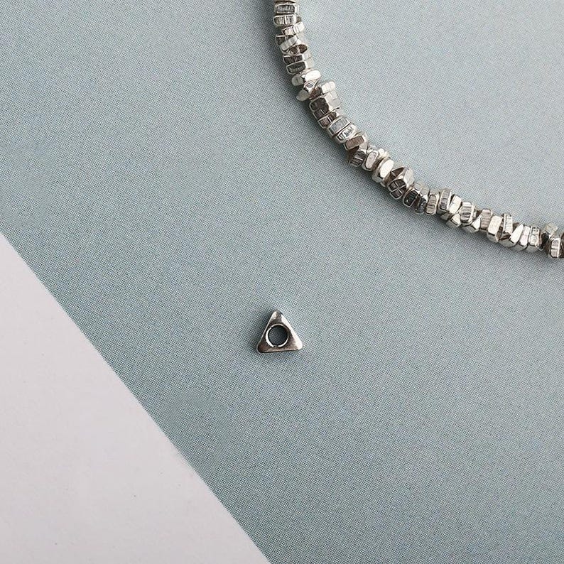 May include: A small, silver-colored triangular charm with a circular hole at its center. The charm is next to a silver beaded necklace. The background is a light blue-gray color.
