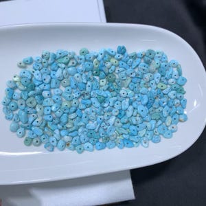 May include: A collection of small, irregular, tumbled turquoise-colored beads with central drill holes. The beads display various shades of light blue, cyan, and pale green, featuring natural textures and smooth, rounded edges throughout the group.