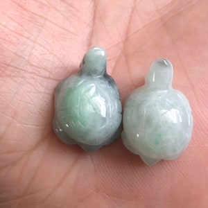 May include: Two jade green turtle charms, one slightly larger than the other, with detailed carvings of the turtle shell. The charms are smooth and polished.