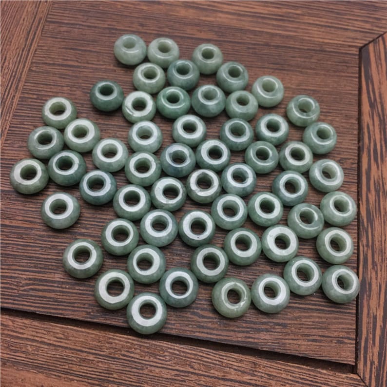 May include: A collection of light green jade beads with a hole in the center of each bead. The beads are arranged in a random pattern on a wooden surface.