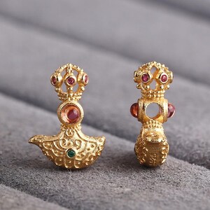 Può includere: Due orecchini color oro con disegni intricati e pietre preziose incastonate. Ogni orecchino presenta una parte superiore a forma di corona con piccole gemme rosse, una gemma rossa centrale e una base decorativa.