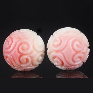 Può includere: Due perle sferiche intagliate in materiale rosa pallido, ciascuna caratterizzata da un motivo a spirale in rilievo che si ripete sulla superficie. Le perle hanno una finitura liscia e lucida con un piccolo foro visibile sulla sommità.