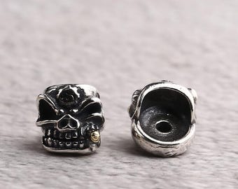 Puede incluir: Dos cuentas de calavera plateadas. Las calaveras tienen detalles, incluyendo cuencas de los ojos, una cavidad nasal y dientes. Una calavera tiene un pequeño acento dorado en un diente. Las cuentas son probablemente para hacer joyas.