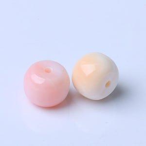 Puede incluir: Dos cuentas redondas, una rosa claro y otra color melocotón cremoso, se muestran sobre un fondo blanco. Cada cuenta tiene un agujero que atraviesa el centro, lo que sugiere que son para la fabricación de joyas o manualidades.