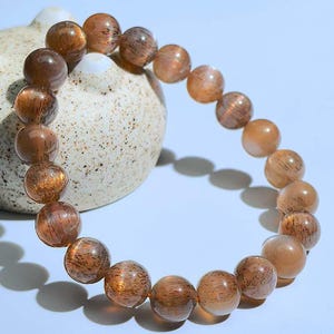 Puede incluir: Un brazalete de cuentas redondas y translúcidas en tonos marrón y ámbar. Las cuentas tienen un aspecto moteado y están ensartadas para formar un círculo completo. El brazalete se exhibe sobre una superficie azul claro. Cada cuenta mide aproximadamente 1 cm de diámetro.