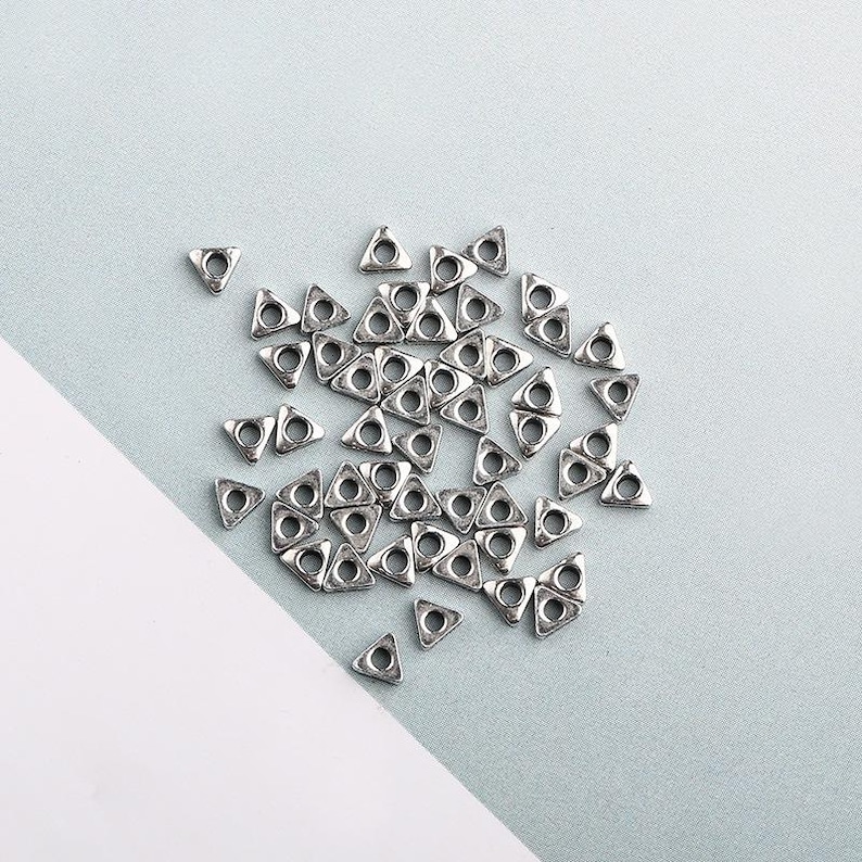 May include: A collection of small, silver-colored metal triangle beads with a hole in the center. The beads are scattered on a light blue and white surface. These beads are ideal for jewelry making and crafting.