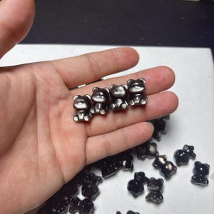 May include: A collection of small, dark gray bear figurines. The bears are sitting upright with rounded features. Some of the bears are held in a hand, while others are scattered on a white surface. The bears have a metallic sheen.