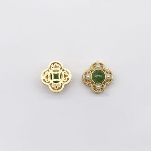 May include: Two gold-colored clover-shaped earrings. One earring features a green stone center and small white pearls. The other earring has a green stone center. Both earrings have a detailed design.