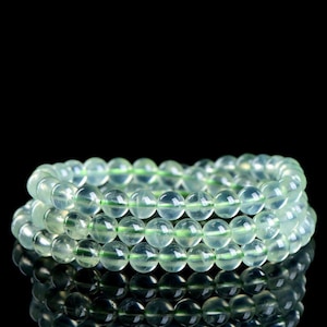May include: A triple-strand bracelet composed of smooth, translucent, pale green spherical beads. The beads have a polished finish and are strung together in three distinct, parallel circular rows, creating a layered appearance.