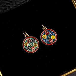 Puede incluir: Dos colgantes de oro con un dise&ntilde;o circular. Cada colgante presenta un mandala colorido con un fondo azul, verde, rojo y amarillo. El mandala tiene una forma de flor con seis p&eacute;talos. Cada p&eacute;talo tiene un s&iacute;mbolo diferente escrito en oro.