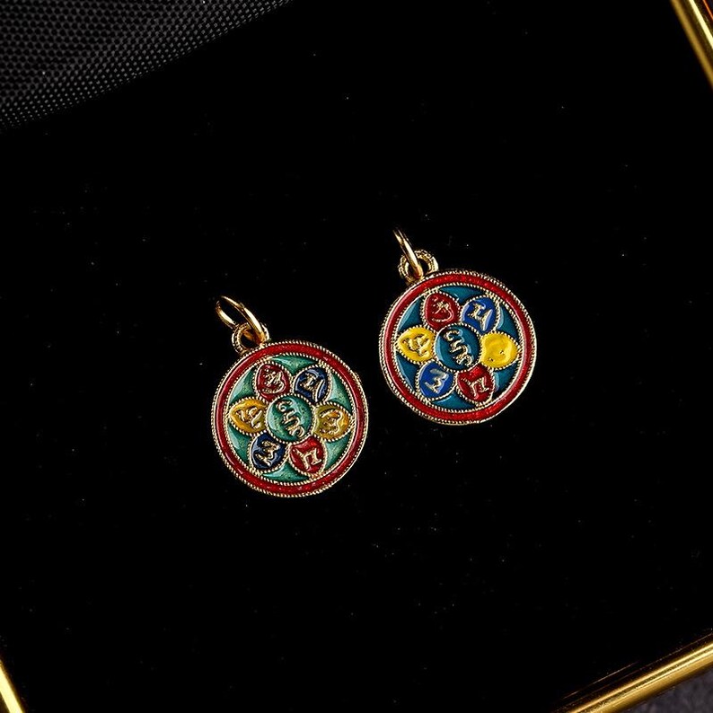 Puede incluir: Dos colgantes de oro con un dise&ntilde;o circular. Cada colgante presenta un mandala colorido con un fondo azul, verde, rojo y amarillo. El mandala tiene una forma de flor con seis p&eacute;talos. Cada p&eacute;talo tiene un s&iacute;mbolo diferente escrito en oro.