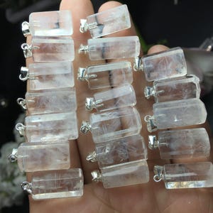 May include: A collection of rectangular, clear crystal pendants with silver-colored bails. The pendants are held in a hand, showcasing their translucent quality and varying internal patterns. Each pendant is approximately 1 inch long.