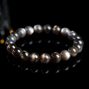 Puede incluir: Un brazalete de cuentas de piedra de sol negra con un borla dorada. Las cuentas son redondas y tienen un acabado brillante e iridiscente.