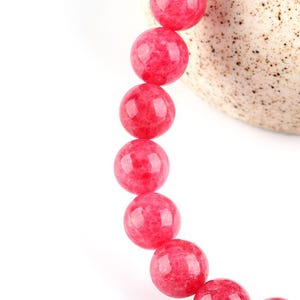 May include: A strand of pink gemstone beads, each bead is round and smooth. The beads are strung together on a clear elastic cord.
