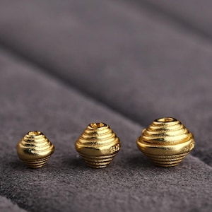 Puede incluir: Tres cuentas de metal de color dorado con un diseño de capas acanaladas. Las cuentas son de diferentes tamaños, siendo la más grande a la derecha y la más pequeña a la izquierda.