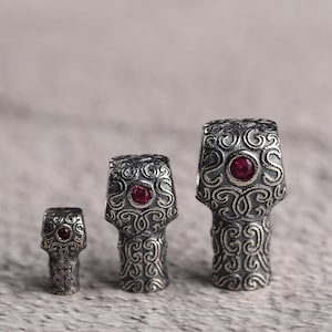 May include: Three silver beads with an intricate scroll design and a central red gemstone. The largest bead is about 2.5 cm tall. The beads have a detailed, vintage appearance, showcasing a classic aesthetic.