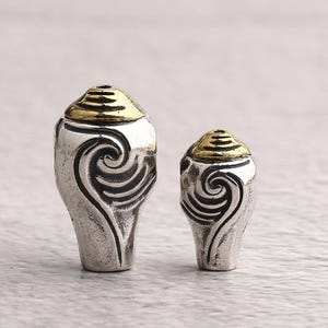 Può includere: Due perline color argento con cime color oro. Ogni perlina presenta un disegno nero a spirale sul davanti. Le perline sono probabilmente per la creazione di gioielli o altri progetti artigianali.