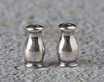 Könnte beinhalten: Zwei kleine, silberfarbene, vasenförmige Perlen. Jede Perle hat eine abgerundete Oberseite mit einer kleinen Öffnung, einen etwas breiteren Körper und einen ausgestellten Sockel. Die Perlen haben ein strukturiertes, leicht angelaufenes Aussehen.