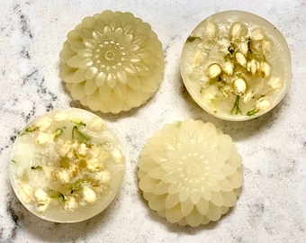 May include: Four round, white, translucent soap bars with floral designs. Two bars have a flower design and two bars have a floral pattern with white flowers and green leaves.