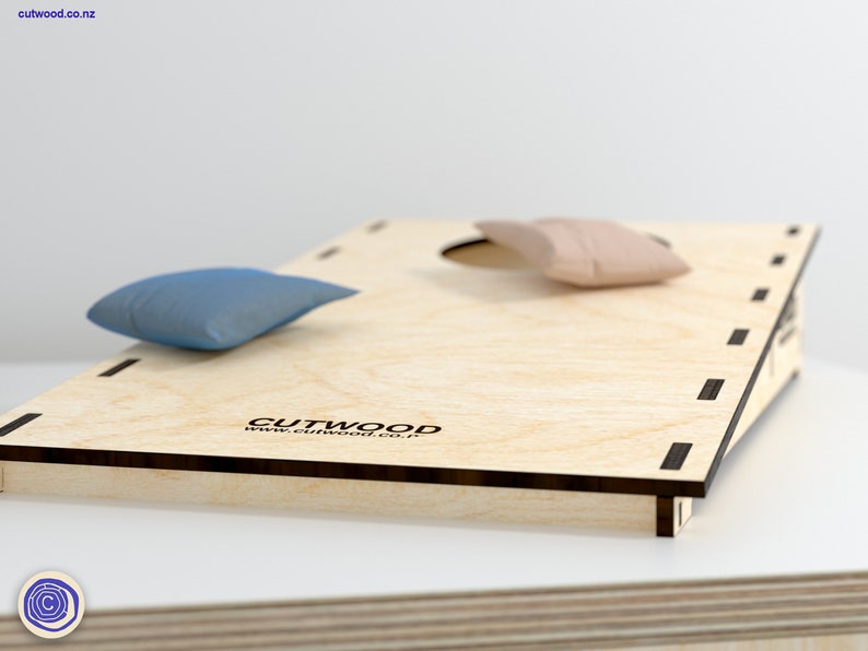 K&ouml;nnte beinhalten: Ein Holz-Beanbag-Wurfspiel mit brauner Beize und schwarzem Besatz. Das Spiel hat zwei Beanbags, einen blauen und einen rosa, und der Text "CUTWOOD www.cutwood.co.nz" ist auf dem Spielbrett aufgedruckt.
