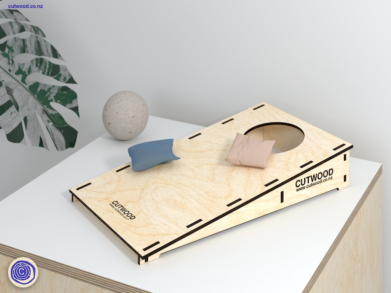 K&ouml;nnte beinhalten: Ein Holz-Beanbag-Wurfspiel mit einer schr&auml;gen Oberfl&auml;che und einem Loch oben. Zwei Beanbags sind enthalten, ein blauer und ein rosa. Das Spiel wird von Cutwood hergestellt und hat deren Website auf der Seite aufgedruckt.