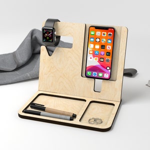 May include: A wooden phone and watch stand with a light wood finish. The stand has a slot for a phone, a watch holder, and two compartments for pens or other small items.