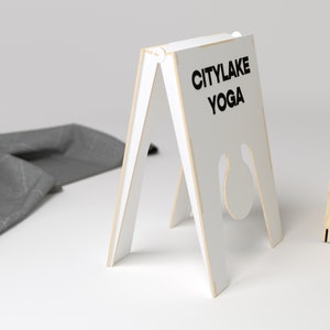 May include: White wooden A-frame sign with the text "CITYLAKE YOGA" in black lettering. The sign is standing on a white surface.