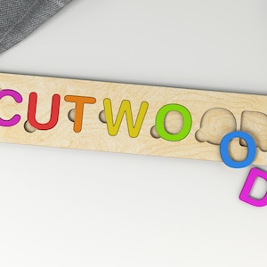 Puede incluir: Un rompecabezas de madera con la palabra "CUTWOOD" escrita en letras de colores. Las letras faltan en sus ranuras y est&aacute;n dispersas alrededor del rompecabezas.