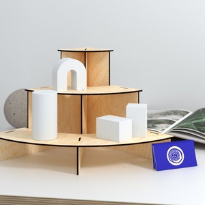 Puede incluir: Un expositor de madera de tres niveles con bordes redondeados. El expositor está hecho de madera clara con ribetes negros. Está decorado con formas geométricas blancas, incluyendo un cilindro, un rectángulo y un arco. Una tarjeta de visita azul con un logotipo blanco está sobre la mesa delante del expositor.