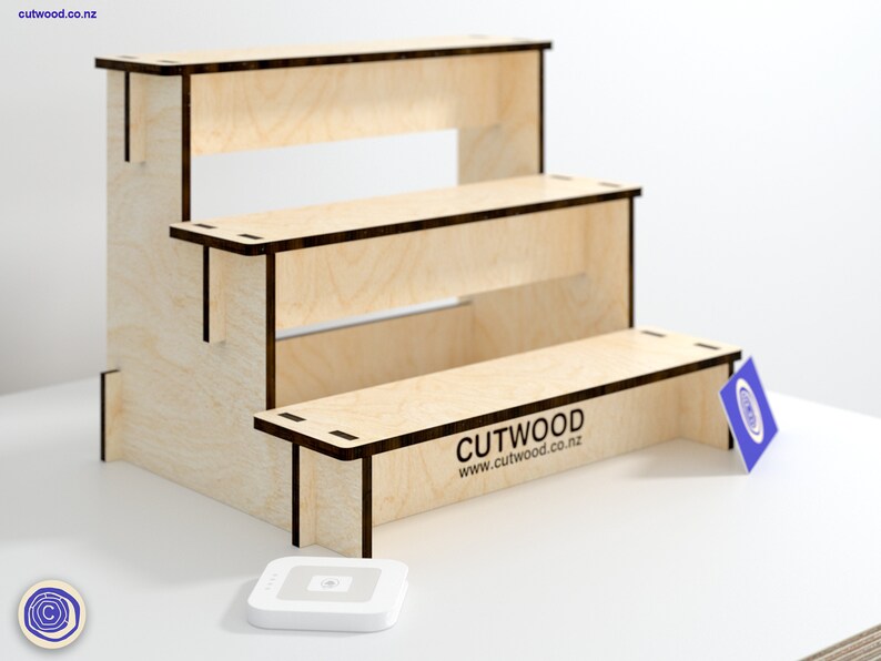 May include: A three-tiered wooden display stand made of light wood with dark brown edges. The stand has a rectangular shape and is about 12 inches tall. The stand is labeled "CUTWOOD www.cutwood.co.nz".