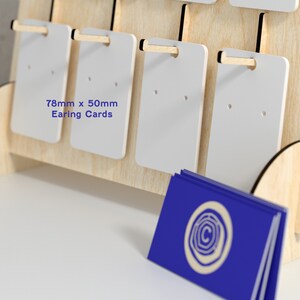May include: A wooden display stand with white earring cards. The cards are 78mm x 50mm and have two holes for displaying earrings. The stand has a blue business card with a logo on it.
