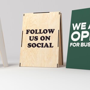 May include: Three wooden A-frame signs. The sign on the left is white with the text "AYLAKE YOGA". The middle sign is made of wood with the text "FOLLOW US ON SOCIAL". The sign on the right is green with the text "WE ARE OPEN FOR BUSINESS".