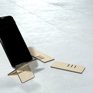 May include: A black smartphone is propped up on a wooden phone stand. The stand is made of three pieces of wood, and the phone is resting on the top piece. The stand is on a white surface.