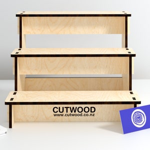 May include: A three-tiered wooden display stand made of light wood with black edges. The stand is empty and ready to display products. The stand is on a white surface. The text "CUTWOOD www.cutwood.co.nz" is printed on the bottom tier.