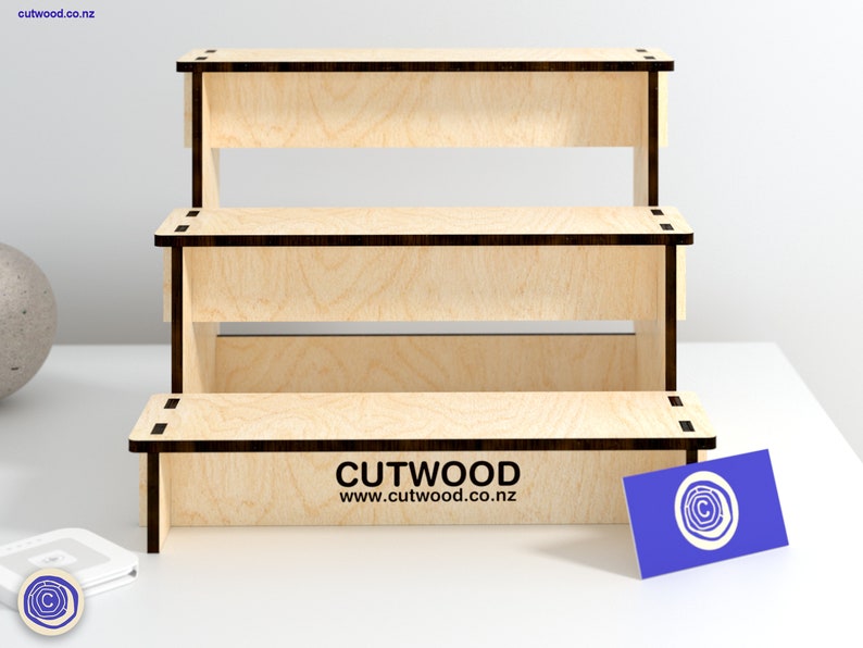May include: A three-tiered wooden display stand made of light wood with black edges. The stand is empty and ready to display products. The stand is on a white surface. The text "CUTWOOD www.cutwood.co.nz" is printed on the bottom tier.