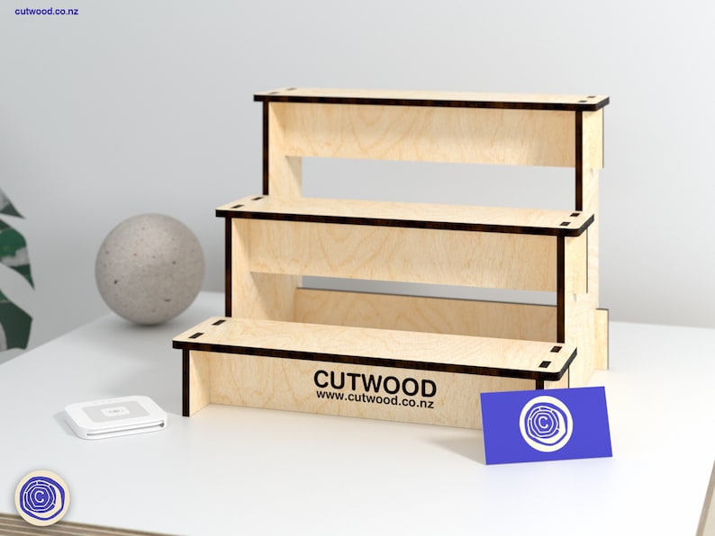 May include: A three-tiered wooden display stand made of light wood with a dark wood trim. The stand is empty and ready to be used to display products. The stand is on a white surface with a business card that says "CUTWOOD www.cutwood.co.nz".