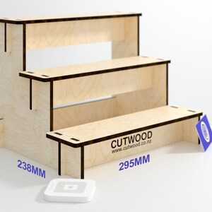May include: A three-tiered wooden display stand made of plywood. The stand is 260mm tall, 238mm wide, and 295mm deep. The stand is made of natural wood with a light brown color. The stand has a simple design with three shelves and a small logo on the bottom shelf that reads "CUTWOOD www.cutwood.co.nz".