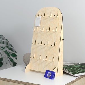 May include: A wooden display stand with a rounded top and multiple hooks for hanging earrings or other small items. The stand is made of light-coloured wood and has a natural finish. It is standing on a white surface.